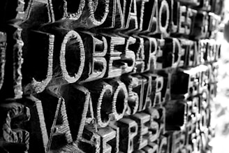 A close-up view of a textured surface featuring intricately carved letters. The lighting casts shadows that enhance the three-dimensional effect of the carving, creating contrast between light and dark areas.