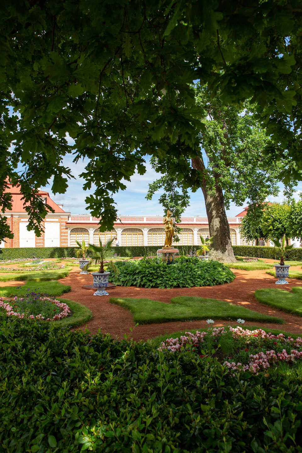 The image depicts a serene garden scene with a large tree casting a dappled shadow on the ground, surrounded by an array of colorful flowers and plants. In the center of the garden is a statue of a person holding a book, adding an artistic touch to the landscape. The garden features a variety of potted plants in different sizes and colors, creating a vibrant and lush atmosphere. The perspective of the image is taken from under the tree's canopy, providing a unique view of the garden below.