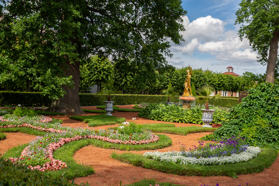 The image depicts a serene garden scene with a large tree at its center, surrounded by an array of colorful flowers and plants. The garden is meticulously arranged into a circular pattern around the tree, creating a sense of harmony and balance. In the background, there are buildings visible through the foliage, adding depth to the scene. The sky above is clear blue with fluffy white clouds, suggesting a bright and sunny day.