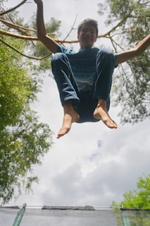 A person is captured in mid-air, likely jumping on a trampoline, surrounded by tall trees. The background shows a cloudy sky with scattered sunlight filtering through the leaves, creating a vibrant aura.