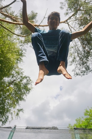 A person is captured in mid-air, likely jumping on a trampoline, surrounded by tall trees. The background shows a cloudy sky with scattered sunlight filtering through the leaves, creating a vibrant aura.
