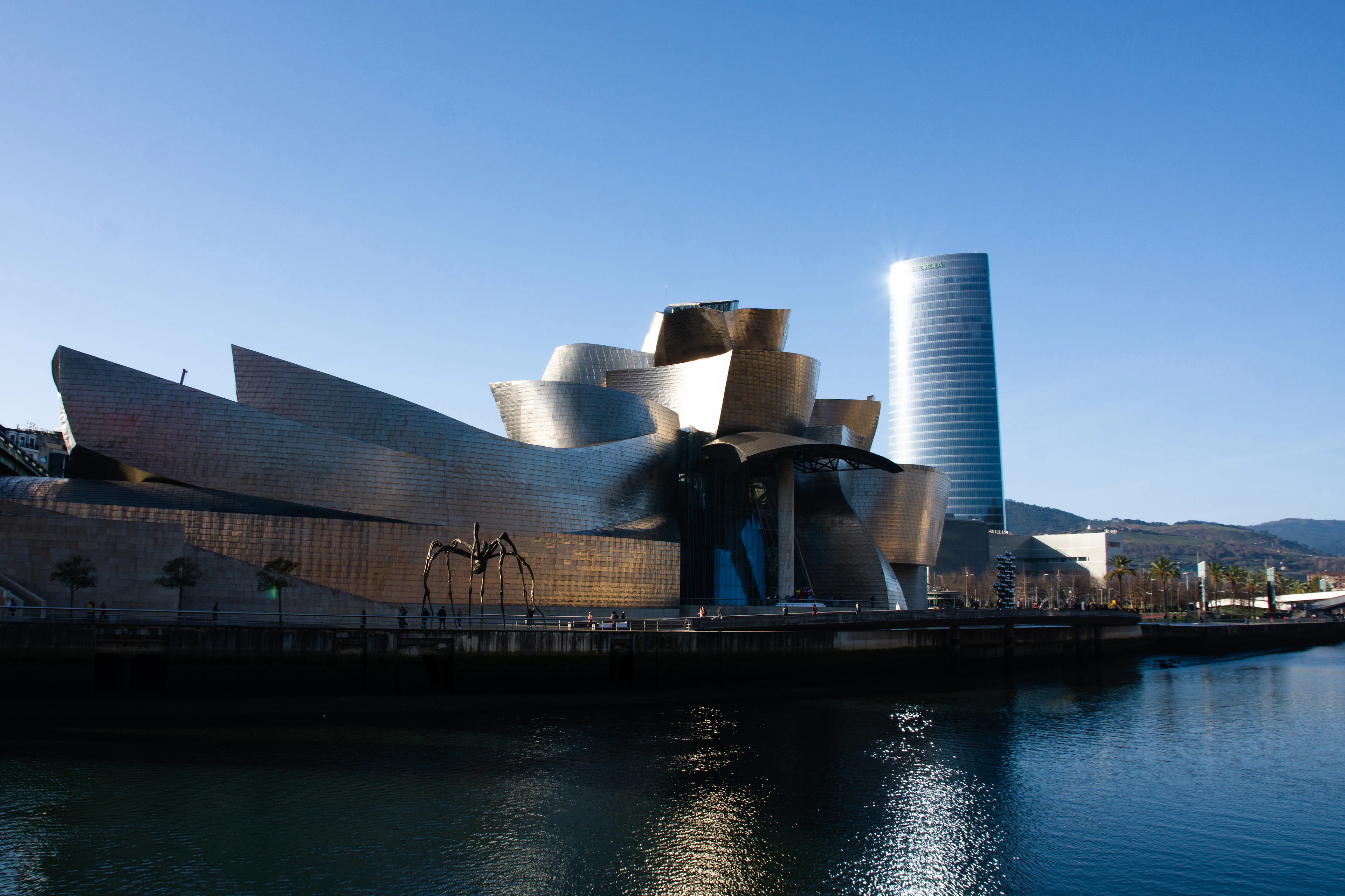The Guggenheim Museum Bilbao in bright daylight, its titanium panels above the Nervión with the Iberdrola Tower alongside