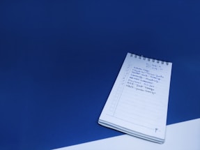 A spiral notebook is placed on a blue surface with a visible checklist handwritten in blue ink. The checklist includes various tasks, some marked with check marks, and dates are noted at the top of the page.