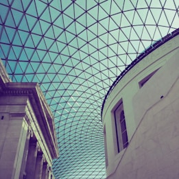 A modern architectural design featuring a large, transparent glass ceiling with a geometric pattern of triangles. Below the ceiling, parts of classical building structures with columns and an arched wall are visible.