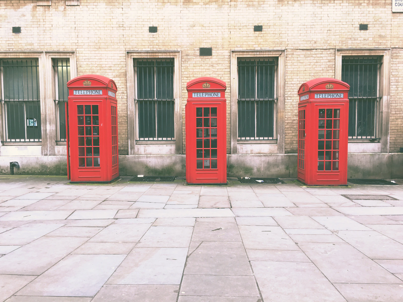 Three red telephone kiosks standing in a row on stone paving in front of a pale brick wall with barred windows