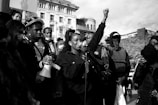 A group of people are gathered in an outdoor setting, with one person at the center speaking into a microphone. Several individuals are holding signs and a megaphone. The central figure has a raised fist, symbolizing solidarity or protest. The background includes buildings and trees, and the crowd displays various expressions of determination and engagement.