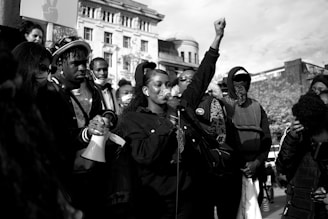 A group of people are gathered in an outdoor setting, with one person at the center speaking into a microphone. Several individuals are holding signs and a megaphone. The central figure has a raised fist, symbolizing solidarity or protest. The background includes buildings and trees, and the crowd displays various expressions of determination and engagement.