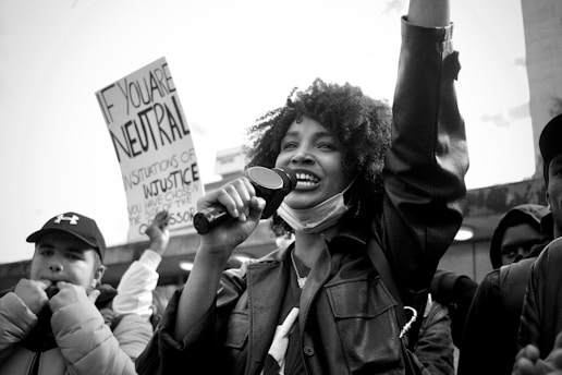 A black and white photograph of a protest scene, where a smiling individual is holding a microphone and raising one arm. This person is surrounded by other protesters, some holding signs, with text partially visible. The crowd appears engaged and passionate.