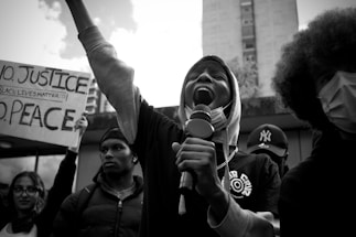 A group of people at a protest. One person in the center is passionately speaking into a microphone while raising one arm. Another person holds a sign that reads 'NO JUSTICE NO PEACE BLACK LIVES MATTER' in the background. The people display emotions of determination and solidarity.
