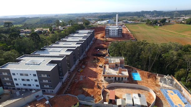 A construction site with several large, modern-looking residential buildings. The landscape in the background shows green fields and distant settlements. There is a swimming pool and a basketball court within the complex. Trees line the edges of the site.