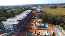 A construction site with several large, modern-looking residential buildings. The landscape in the background shows green fields and distant settlements. There is a swimming pool and a basketball court within the complex. Trees line the edges of the site.