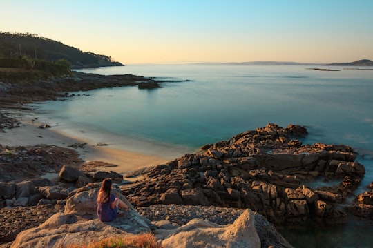 woman in blue and red dress sitting on rock near body of water during daytime in Ría de Aldán Spain