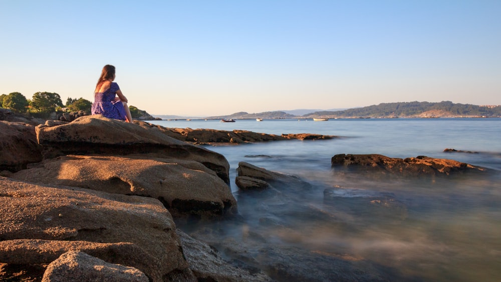 mulher na jaqueta cor-de-rosa sentada na rocha perto do corpo de água durante o dia