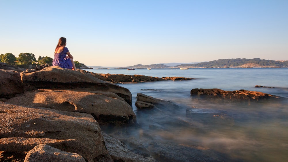 woman in pink jacket sitting on rock near body of water during daytime