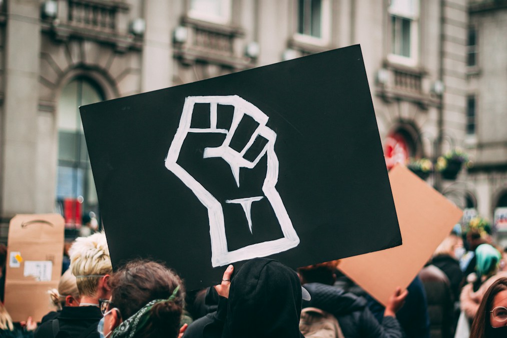 Pancarte noire ornée d’un poing levé dessiné à la main, brandie au-dessus d’une foule de manifestants (photo d’illustration)