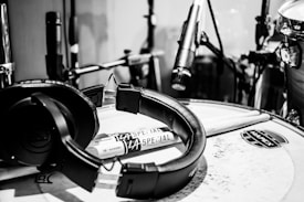 A monochrome image featuring a drum set with a focus on a pair of headphones resting on a snare drum. Drumsticks are laid across the drum, and several microphones are positioned around the set for recording. Visible branding includes 'LA Special' on the drumsticks and 'Mapex' on the drum.