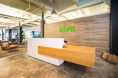 A modern office reception area features a sleek, minimalist wooden desk with a white base against a wood-paneled wall. The word 'kiva' is displayed in bright green on the wall. There are hanging lights and a polished concrete floor. To the left, several wooden chairs with cushions and plants enhance the contemporary office environment.