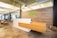 A modern office reception area features a sleek, minimalist wooden desk with a white base against a wood-paneled wall. The word 'kiva' is displayed in bright green on the wall. There are hanging lights and a polished concrete floor. To the left, several wooden chairs with cushions and plants enhance the contemporary office environment.