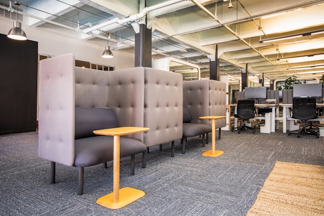A modern, open office space featuring high-backed lounge chairs with small wooden side tables. The chairs are upholstered in a light gray fabric and provide a semi-private seating area. In the background, there are workstations with computers and black office chairs. The ceiling showcases exposed ductwork and lighting fixtures, enhancing the contemporary industrial aesthetic.