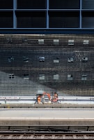 A construction scene near a train platform, featuring workers in orange safety gear operating machinery below a large, reflective grid-like structure.