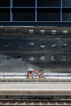 A construction scene near a train platform, featuring workers in orange safety gear operating machinery below a large, reflective grid-like structure.