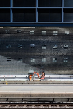 A construction scene near a train platform, featuring workers in orange safety gear operating machinery below a large, reflective grid-like structure.