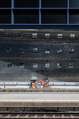 A construction scene near a train platform, featuring workers in orange safety gear operating machinery below a large, reflective grid-like structure.
