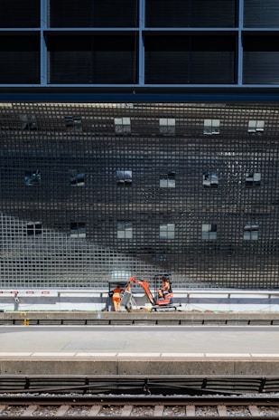 A construction scene near a train platform, featuring workers in orange safety gear operating machinery below a large, reflective grid-like structure.