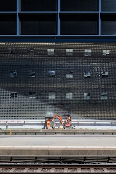A construction scene near a train platform, featuring workers in orange safety gear operating machinery below a large, reflective grid-like structure.