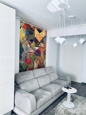 A modern living room with a large gray sofa placed against a wall. Above the sofa, there is a vibrant, multicolored geometric pattern artwork. A white, round coffee table is positioned in front of the sofa with a couple of remote controls on it. The room features minimalist lighting with several white pendant lights hanging above and a light gray rug covering part of the floor.