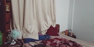 A bedroom scene with a neatly made bed covered with a patterned bedspread and several pillows. There is a nightstand on the right side of the bed with various items on it. The headboard is flanked by tall beige curtains. A small desk fan, a green water bottle, and shelves containing various items are visible on the left side.