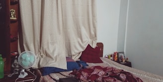 A bedroom scene with a neatly made bed covered with a patterned bedspread and several pillows. There is a nightstand on the right side of the bed with various items on it. The headboard is flanked by tall beige curtains. A small desk fan, a green water bottle, and shelves containing various items are visible on the left side.