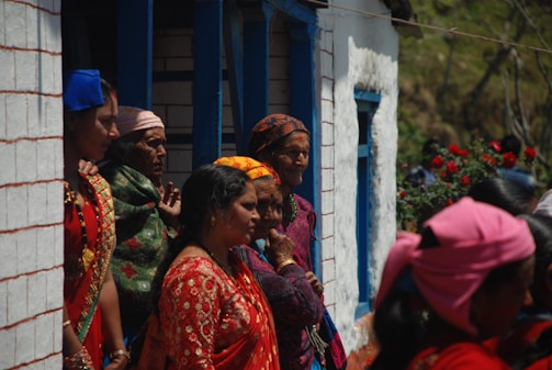 A group of women dressed in vibrant traditional clothing stand outside a building with white and blue accents. The women appear to be engaged in conversation or observing something out of view. The scene includes red flowers blooming nearby, adding a pop of color to the setting.