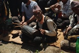 A group of people sitting on a mat outdoors, engaged in playing traditional musical instruments. The central figure, an elderly man, is playing a drum, while others are playing flutes and other percussion instruments. They are wearing traditional hats and attire.