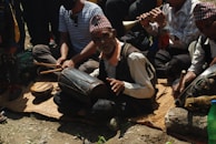 A group of people sitting on a mat outdoors, engaged in playing traditional musical instruments. The central figure, an elderly man, is playing a drum, while others are playing flutes and other percussion instruments. They are wearing traditional hats and attire.