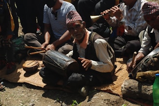 A group of people sitting on a mat outdoors, engaged in playing traditional musical instruments. The central figure, an elderly man, is playing a drum, while others are playing flutes and other percussion instruments. They are wearing traditional hats and attire.
