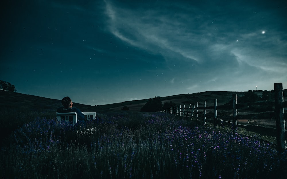 homme en veste noire assis sur une clôture en bois marron sous le ciel bleu pendant la nuit
