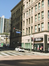 The image depicts an urban street corner with a historic multi-story building featuring ornate architecture and large windows. A T-Mobile store is situated on the ground floor of this building beside a Broadway street sign. A white car is parked on the side of the road, and a pedestrian walks nearby under a clear blue sky.