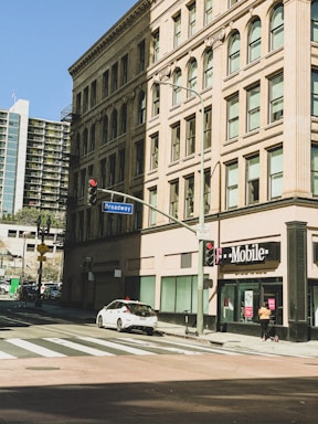 The image depicts an urban street corner with a historic multi-story building featuring ornate architecture and large windows. A T-Mobile store is situated on the ground floor of this building beside a Broadway street sign. A white car is parked on the side of the road, and a pedestrian walks nearby under a clear blue sky.