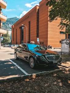 A sleek black Mercedes parked in a shaded area with a mountainous backdrop. It is positioned near a tall, rust-colored brick building under a clear blue sky. The setting appears urban with other cars and trees visible nearby.