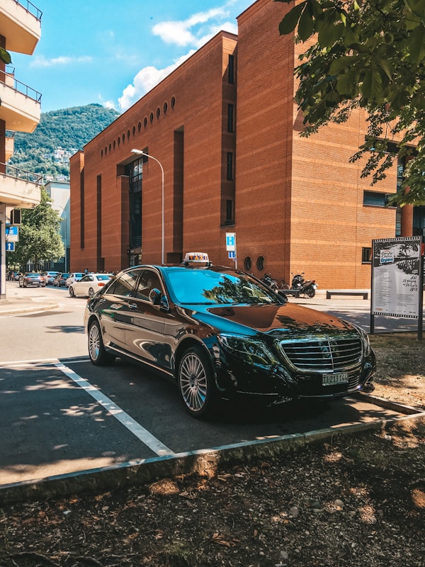 A sleek black Mercedes parked in a shaded area with a mountainous backdrop. It is positioned near a tall, rust-colored brick building under a clear blue sky. The setting appears urban with other cars and trees visible nearby.