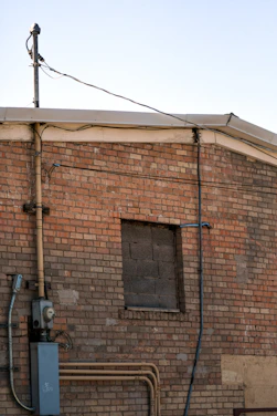 A brick wall with an old, blocked-up window and various exposed utility cables and pipes. The wall appears aged, with a mix of red and beige bricks. An electrical meter box and a conduit pipe are attached to the wall.