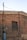 A brick wall with an old, blocked-up window and various exposed utility cables and pipes. The wall appears aged, with a mix of red and beige bricks. An electrical meter box and a conduit pipe are attached to the wall.