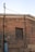 A brick wall with an old, blocked-up window and various exposed utility cables and pipes. The wall appears aged, with a mix of red and beige bricks. An electrical meter box and a conduit pipe are attached to the wall.