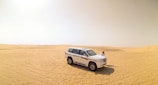 A white SUV is parked on the vast, golden sand dunes under a clear sky. The scene captures the feeling of isolation and adventure, with a person standing beside the vehicle, dressed in traditional attire, seemingly taking in the expansive desert landscape.