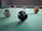 Several billiard balls are placed on a green pool table surface, with the black 8-ball prominently in the foreground. The table is situated in a dimly lit room, creating a subtle focus on the billiard balls.
