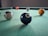 Several billiard balls are placed on a green pool table surface, with the black 8-ball prominently in the foreground. The table is situated in a dimly lit room, creating a subtle focus on the billiard balls.