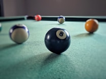 Several billiard balls are placed on a green pool table surface, with the black 8-ball prominently in the foreground. The table is situated in a dimly lit room, creating a subtle focus on the billiard balls.