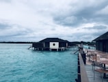 A tranquil scene featuring overwater villas on stilts, set against a backdrop of a cloudy sky. The water is a vibrant turquoise, and there are several wooden tables and chairs on a nearby deck, suggesting a place for relaxation.