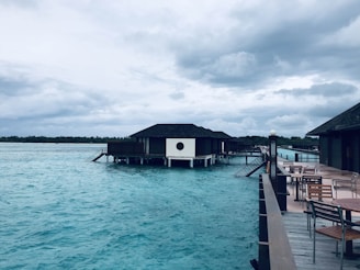 A tranquil scene featuring overwater villas on stilts, set against a backdrop of a cloudy sky. The water is a vibrant turquoise, and there are several wooden tables and chairs on a nearby deck, suggesting a place for relaxation.